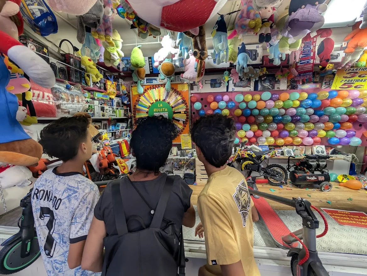 Interior de una barraca de feria vista desde el mostrador: decenas de peluches colgando del techo, estanterías con cajas de consolas y electrónica, un panel circular con el rótulo «La Ruleta», una pared cubierta de globos de colores y, al fondo, una moto infantil y un patinete entre los premios