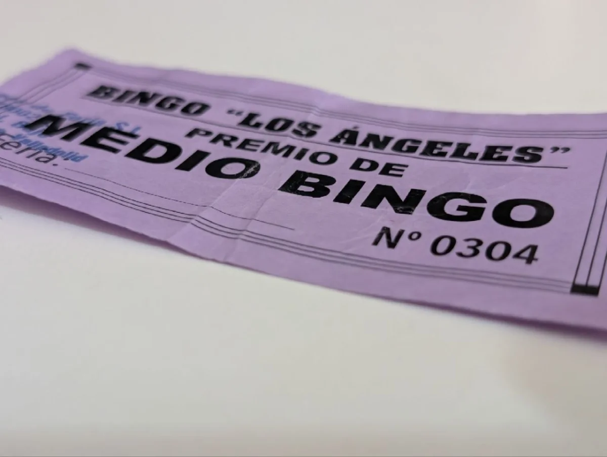 Billete de feria de papel malva, arrugado, sobre una mesa blanca. En letras negras muy grandes se lee «Bingo Los Ángeles · Premio de MEDIO BINGO» y, debajo, un número de serie impreso: Nº 0304