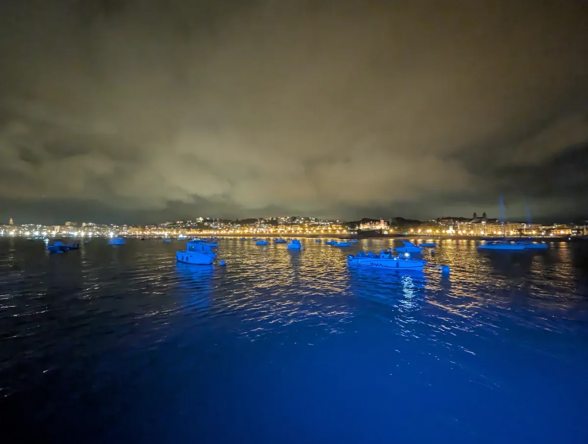 Decenas de embarcaciones fondeadas en la bahía de la Concha a la hora azul, iluminadas de azul por la luz de cubierta, con el paseo y la ciudad encendidos al fondo