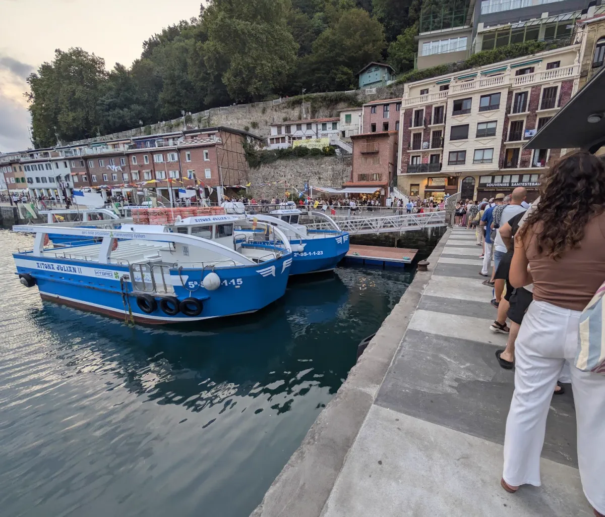Dos motoras azules amarradas en el muelle de Donostia al atardecer, con una fila de personas esperando en el borde del muelle para embarcar