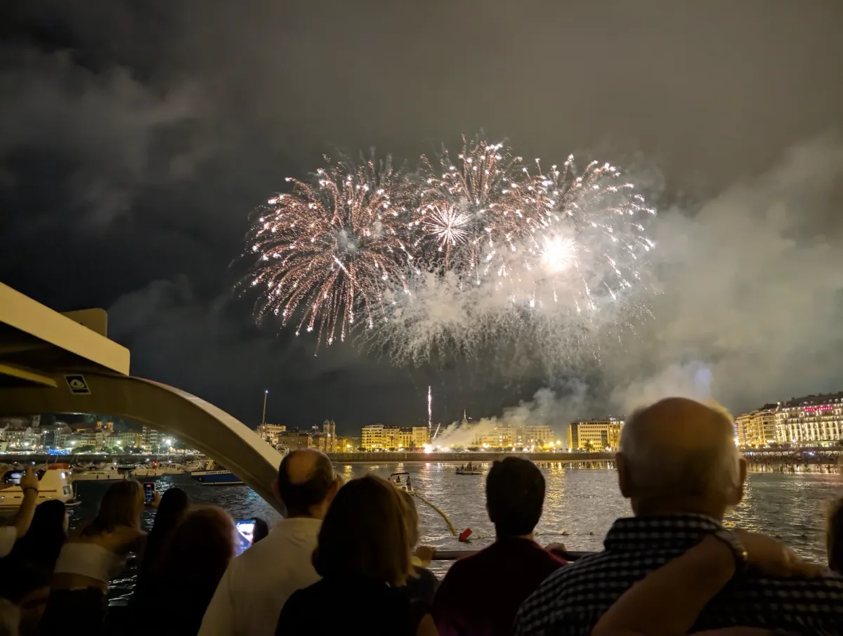 Varios ramilletes de fuegos artificiales abriéndose sobre la bahía de la Concha, vistos por encima de las cabezas del pasaje de una motora, con la ciudad iluminada al fondo