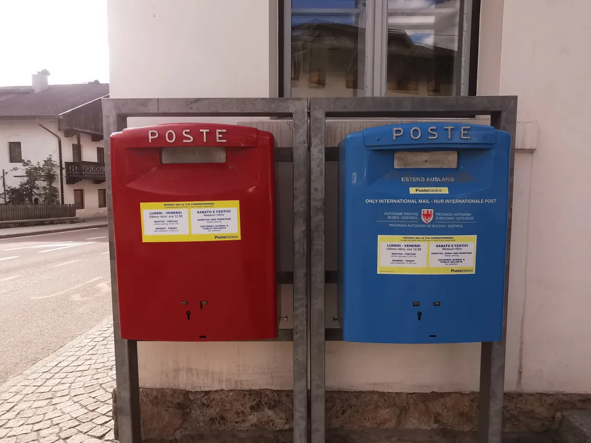 Dos buzones de correos de Poste Italiane montados en un bastidor metálico junto a una fachada: uno rojo y otro azul para el correo internacional, este con el escudo de la provincia autónoma de Bolzano