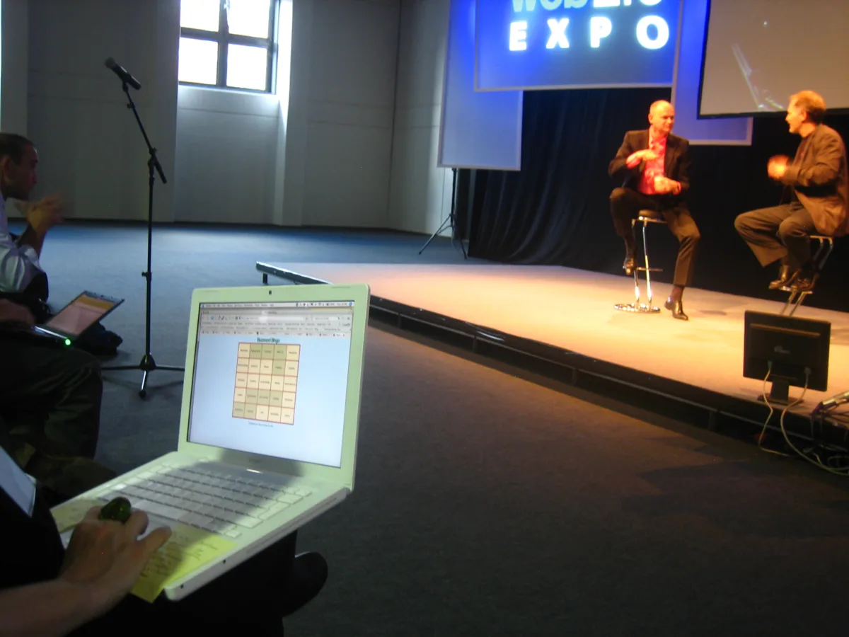 Sala de conferencias: al fondo, dos ponentes sentados en taburetes bajo un rótulo luminoso que dice WEB 2.0 EXPO; en primer plano, un portátil abierto con un cartón de bingo en el navegador