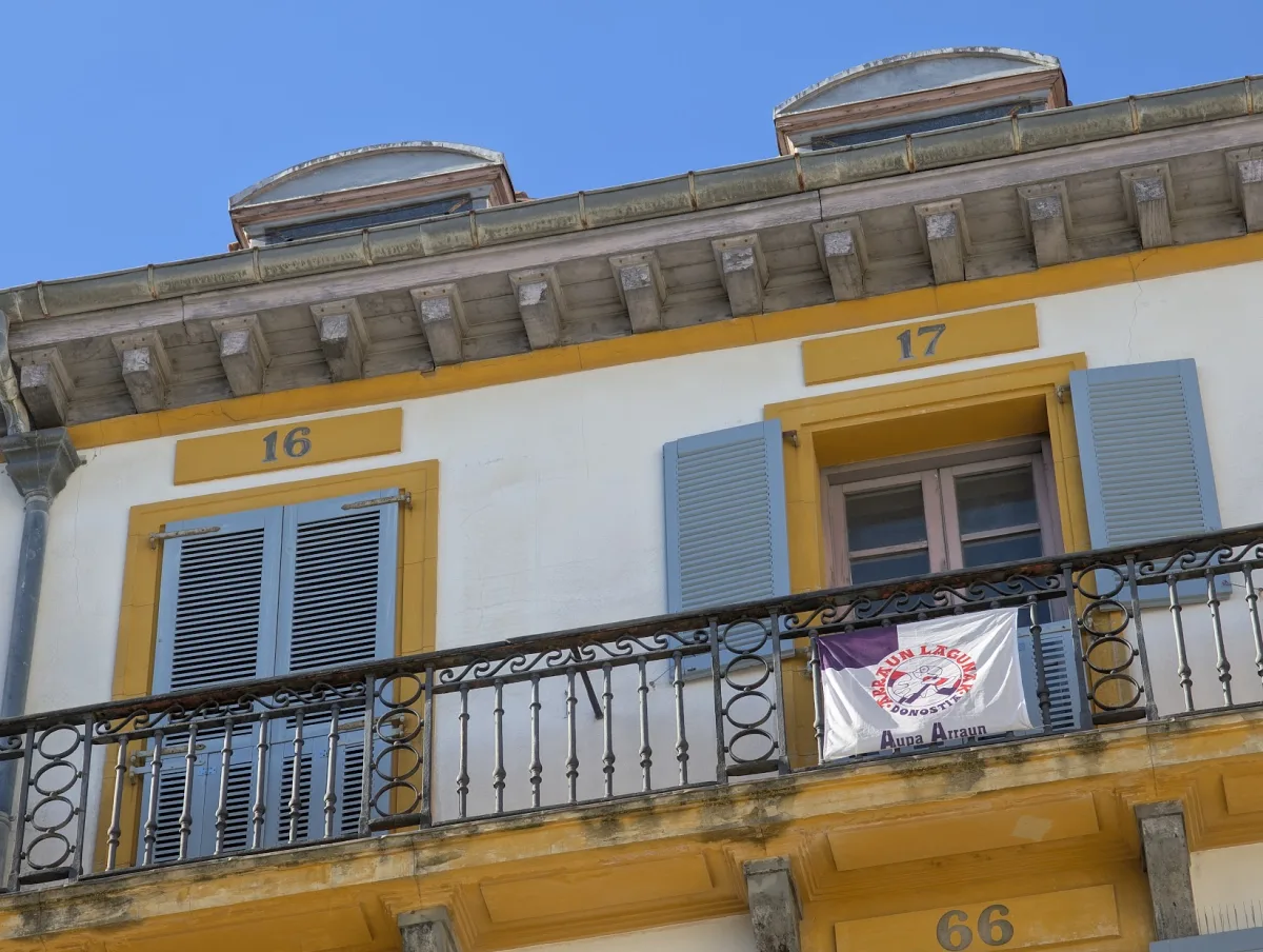 Balcón de hierro de la plaza de la Constitución, con los números 16 y 17 pintados sobre las ventanas, del que cuelga una bandera de Arraun Lagunak Donostia con el lema «Aupa Arraun»