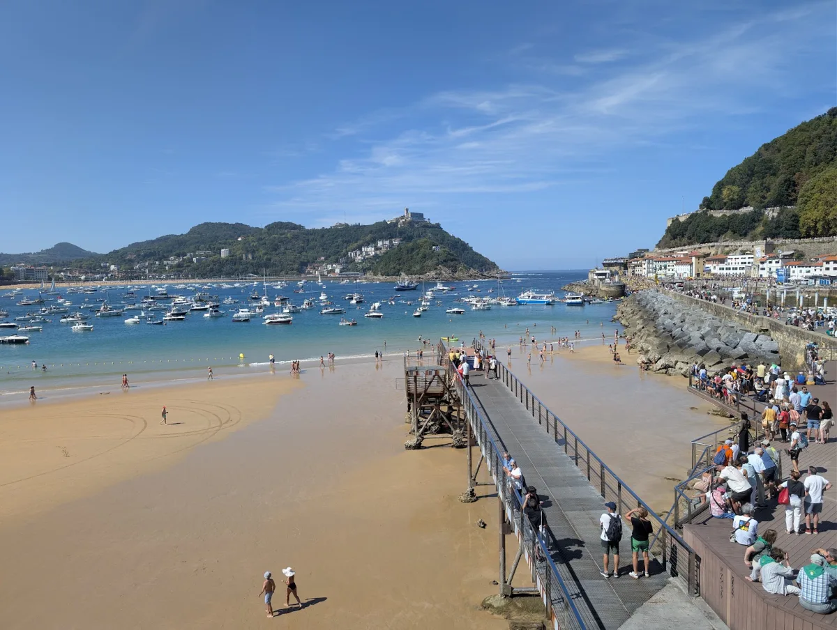 La bahía de la Concha vista desde lo alto del Club Náutico en un día soleado, con la playa en marea baja, la pasarela, decenas de barcos fondeados, el monte Igueldo al fondo y el paseo del puerto lleno de gente a la derecha
