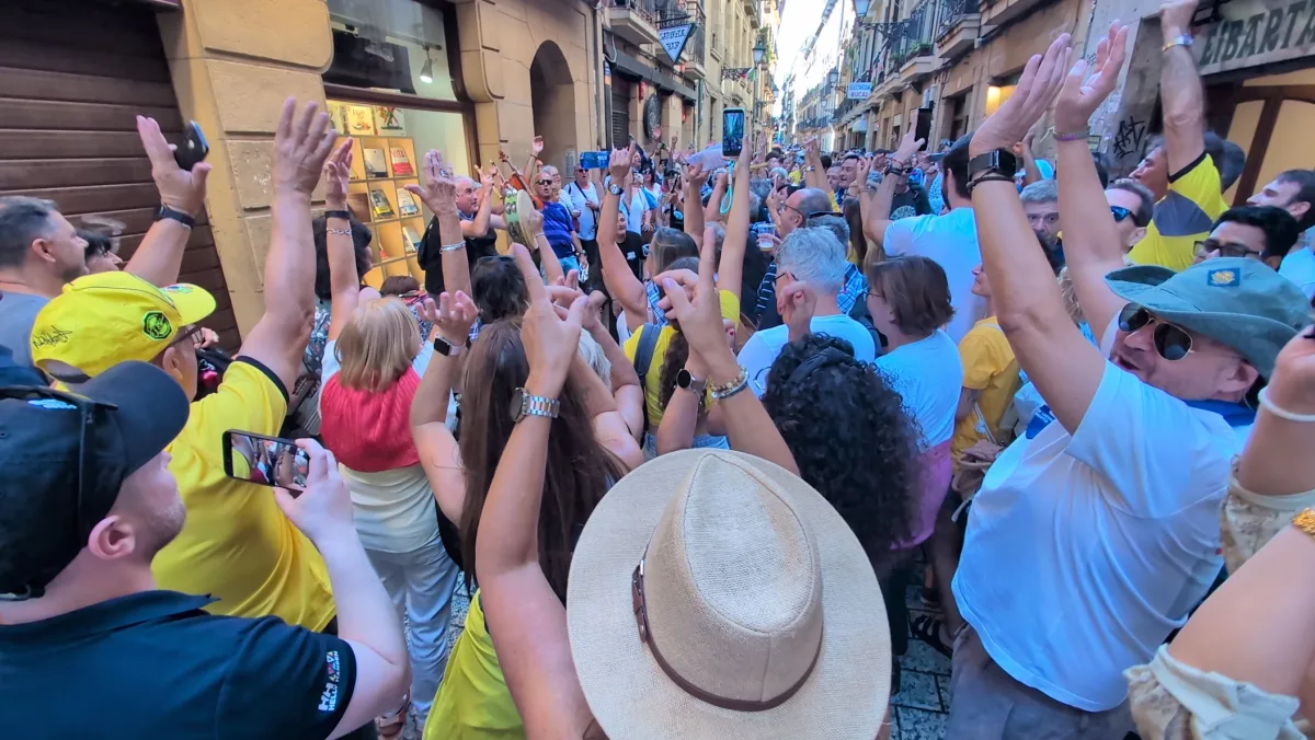 Una calle estrecha de la Parte Vieja de Donostia abarrotada de gente con los brazos en alto alrededor de un grupo de música, con camisetas y gorras amarillas de Orio mezcladas con otros colores y varios móviles grabando