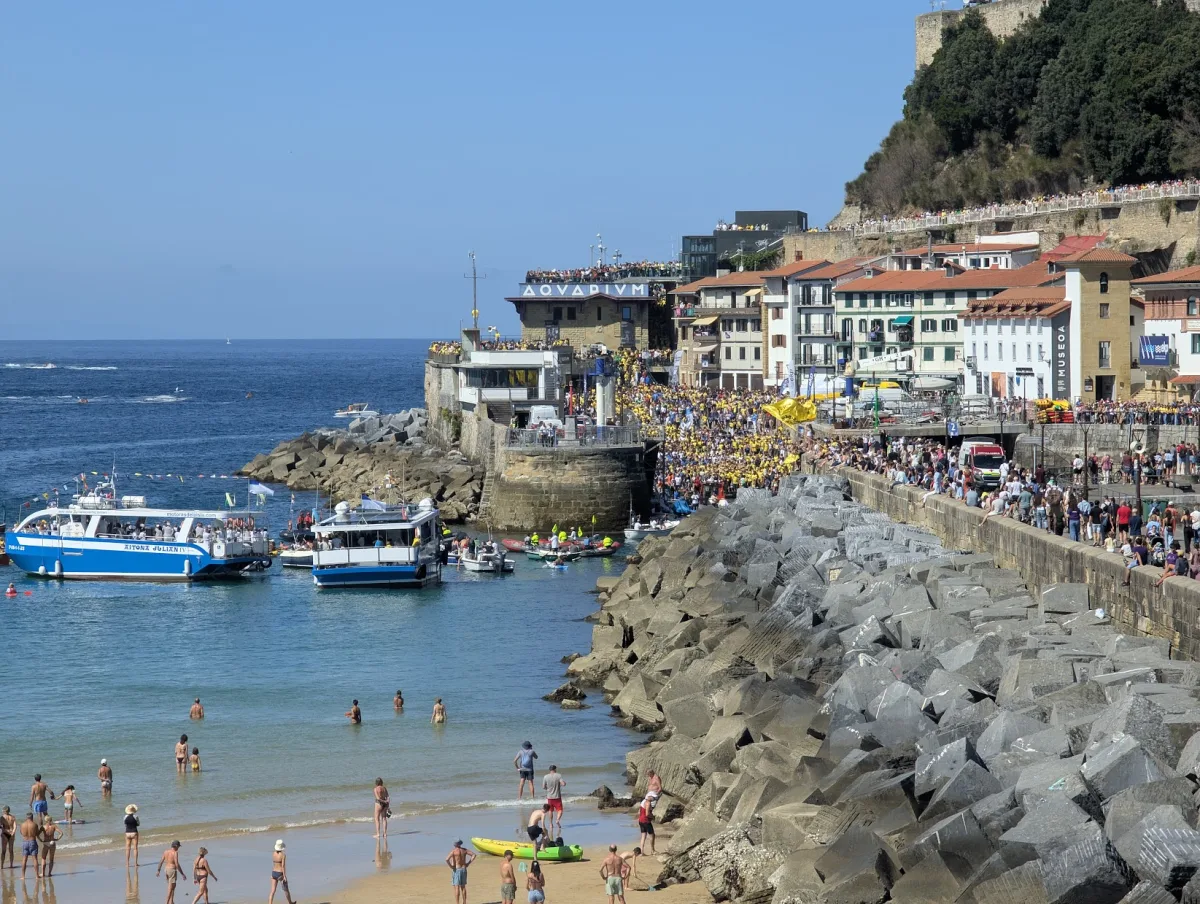 El puerto de Donostia abarrotado de aficionados vestidos de amarillo junto al Aquarium, con el paseo de Urgull lleno de gente en lo alto, dos motoras azules en el agua y bañistas en la playa en primer plano