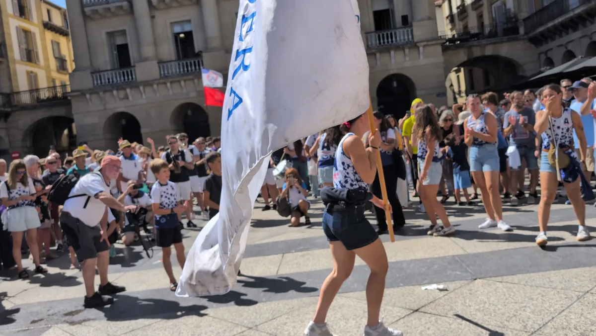 Una remera de Arraun Lagunak, de espaldas, avanza por la plaza de la Constitución con la bandera blanca al hombro, mientras compañeras del equipo y el público aplauden alrededor y una banda toca al fondo