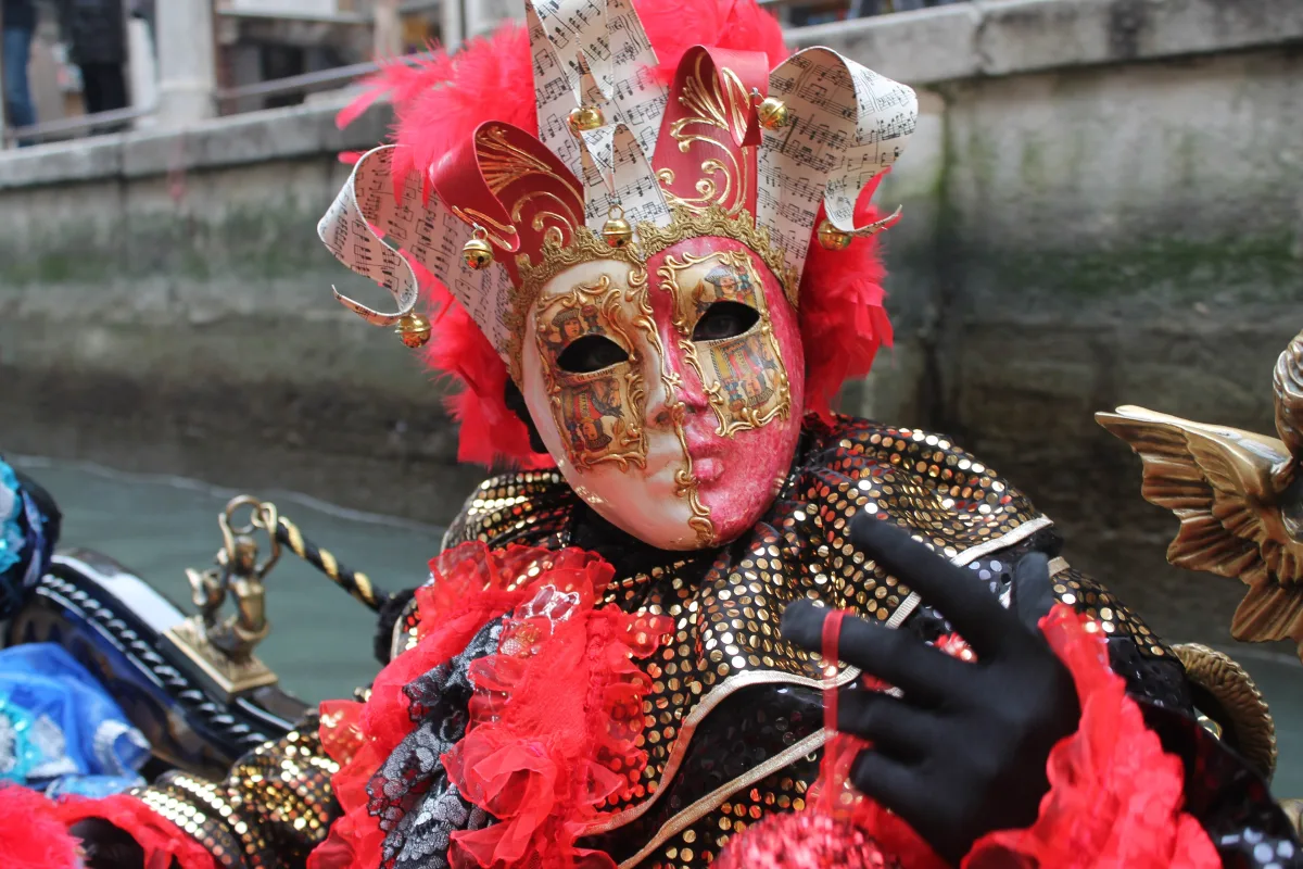 Persona disfrazada para el carnaval de Venecia, con máscara roja, blanca y dorada, tocado de partituras y guantes negros, sentada en una góndola junto a un canal