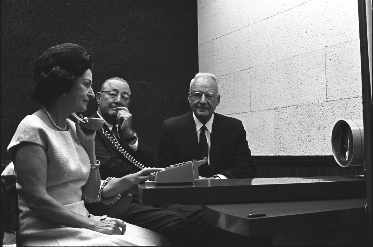 Fotografía en blanco y negro de 1964: una mujer habla por un teléfono junto a un Picturephone mientras dos hombres trajeados sentados a su lado observan