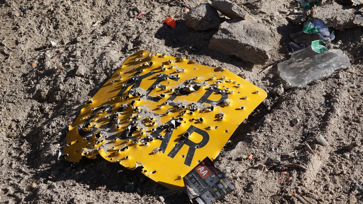 Señal de tráfico amarilla con el rótulo «KEEP CLEAR», completamente acribillada a balazos y tirada boca arriba sobre la tierra, con una caja vacía de cartuchos de escopeta junto a ella