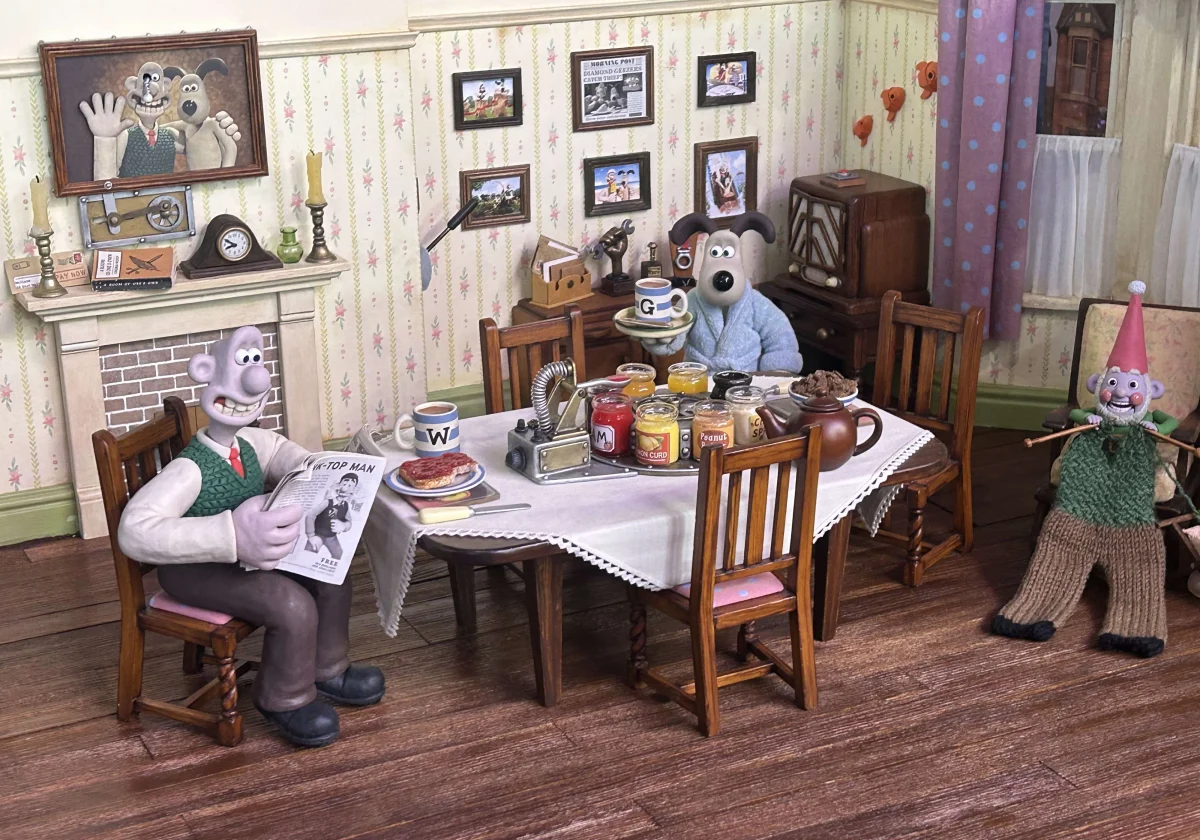 Decorado en miniatura del comedor de Wallace y Gromit: Wallace sentado leyendo una revista, Gromit de pie con una bandeja y una taza, un gnomo de jardín con jersey verde a la derecha, y la pared empapelada llena de cuadros diminutos
