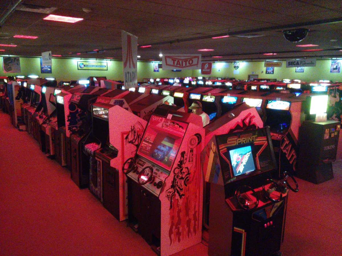 Un salón lleno de muebles recreativos en filas, con las pantallas encendidas y carteles de Atari, Taito, Nintendo y Sega colgados del techo