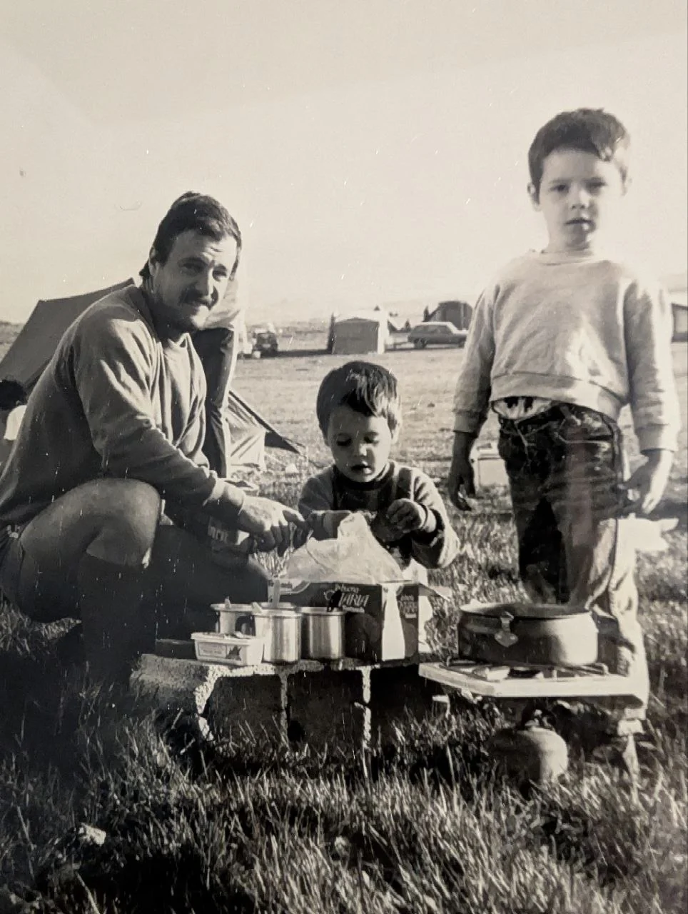 Fotografía antigua en blanco y negro: un hombre con bigote agachado junto a una cocina de camping montada sobre bloques de hormigón, un niño pequeño metiendo las manos en una caja de cartón y otro algo mayor de pie a la derecha, con tiendas de campaña y coches al fondo